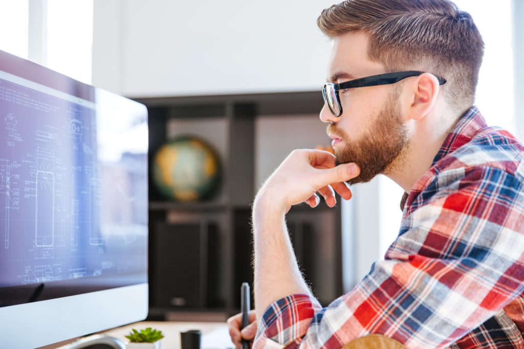 Profile of concentrated bearded man in glasses sitting and drawing blueprints on computer