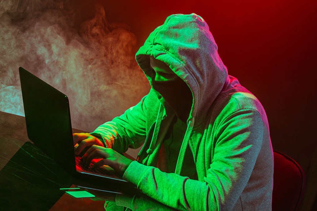 Hooded computer hacker stealing information with laptop on colored studio background