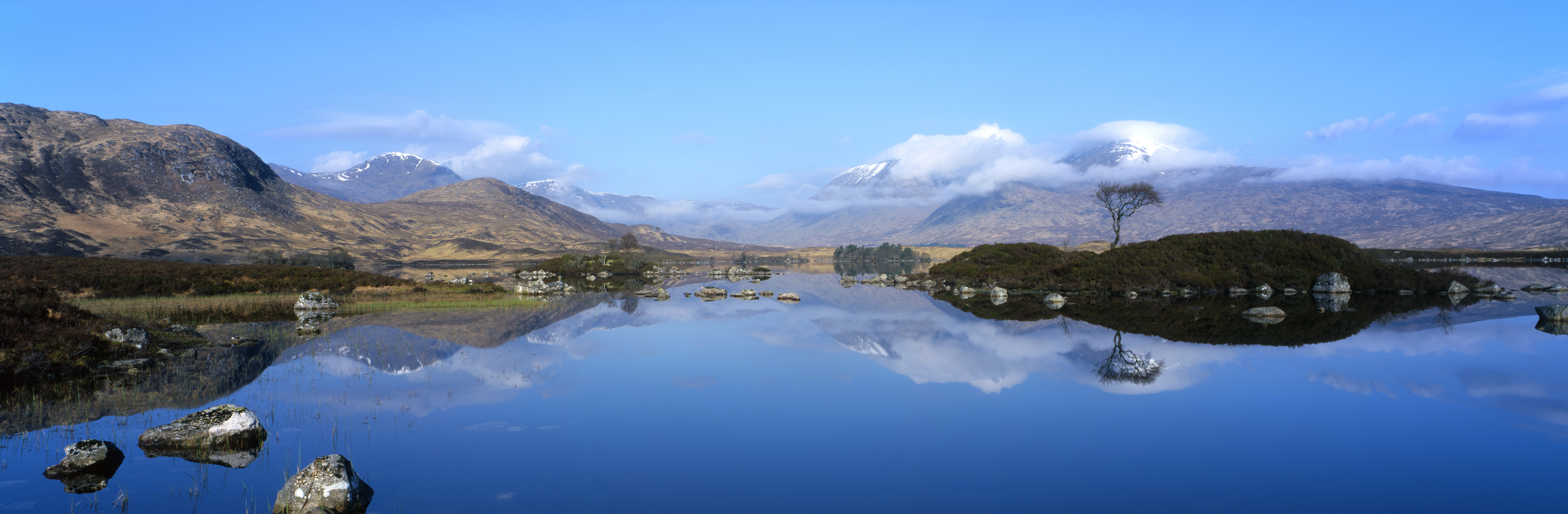 Clouds, mountains, stones and trees mirrored in water. Loch Na-Achlaise, Scotland.