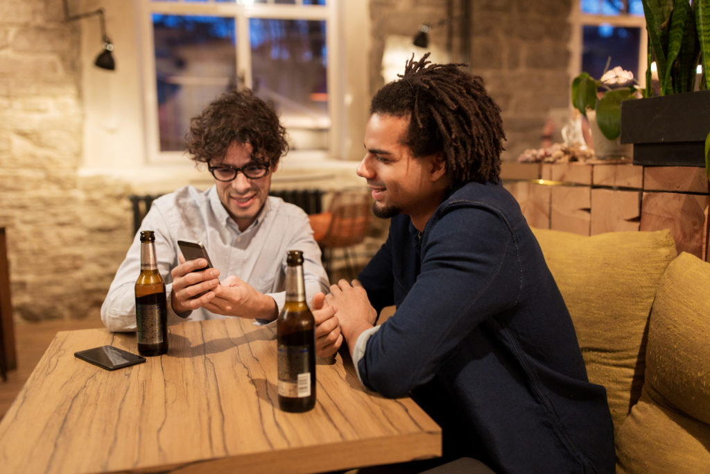 people, men, leisure, friendship and technology concept - happy male friends with smartphones drinking bottled beer at bar or pub